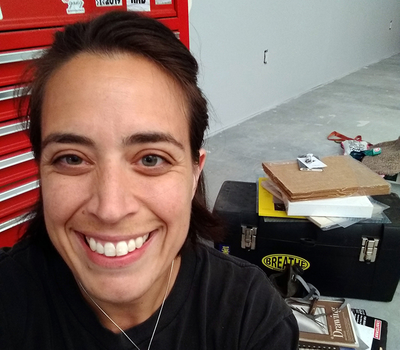 a woman with brown hair smiles at the camera with two toolboxes and art supplies on the floor behind her